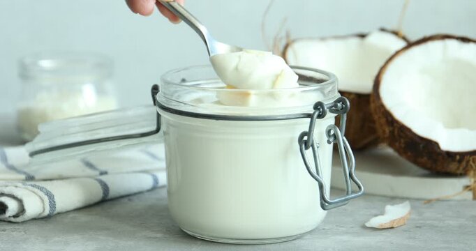 Woman taking delicious coconut yogurt from glass jar with spoon at light grey table, closeup