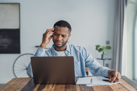 A young man sits at a wooden table in a bright home office, working on a laptop and reviewing papers with a puzzled expression, suggesting concentration, remote work and problem solving.