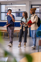 Diverse student friends walking and talking in campus lounge, carrying folders, tablet, headphones © wavebreak3
