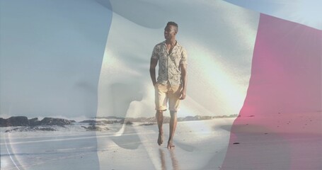 Obraz premium Walking man in patterned short-sleeve shirt, shorts on wet sand, with faint tricolor flag overlay
