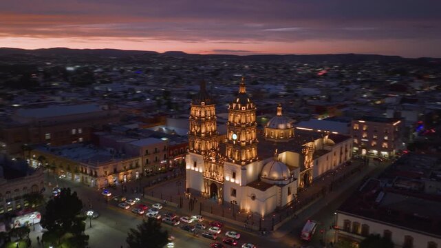 Aerial hyperlapse captures stunning Durango Cathedral bathed in golden lights at twilight in Durango, Mexico. Vibrant pink and purple sunset sky stretches across horizon as city lights twinkle below.