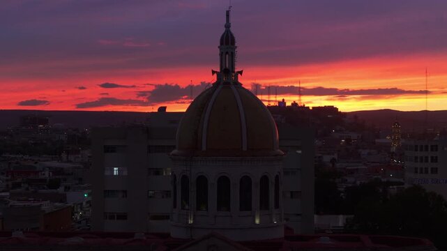 Aerial view of ornate church dome with golden cupola and bell tower silhouetted against dramatic orange and purple sunset sky in Durango Mexico. City buildings and mountains visible in twilight.