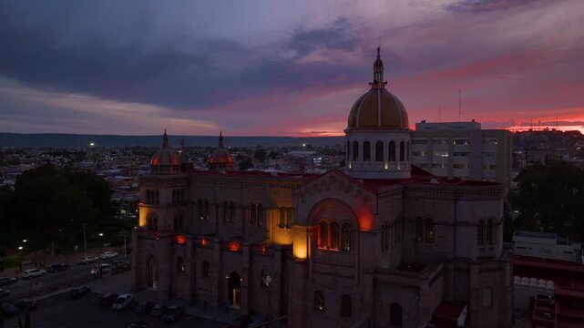 Aerial view of majestic cathedral with golden dome illuminated at twilight in Durango, Mexico. Dramatic purple and orange sunset sky with moody clouds over the historic cityscape as warm lights glow.