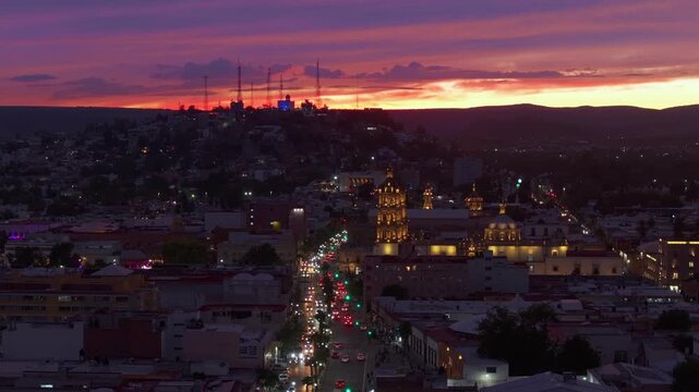 Durango Mexico cityscape at sunset with illuminated historic cathedral dominating the skyline. Traffic flows along main avenue as vibrant orange and purple clouds paint the sky above the colonial.