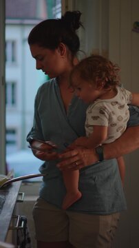 Mother holding toddler on hip while cooking at stove capturing multitasking caregiving routine and everyday domestic life in warm home environment