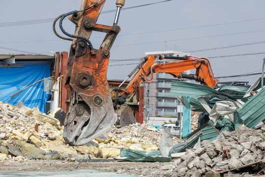 Excavator with heavy-duty demolition crusher works on concrete debris and scrap metal while another excavator in background busily clears rubble with hydraulic arm at demolition site.
