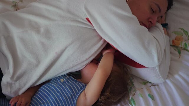 Sleeping mother curled on bed while toddler lies close under her arm capturing protective parenting gesture and calm domestic family moment