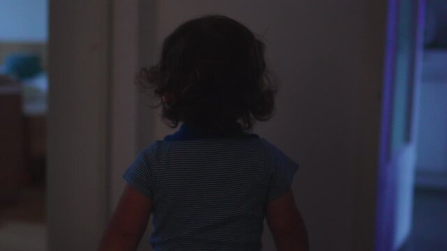 Toddler walking down dim hallway toward lit room deeper inside home, quiet childhood moment expressing curiosity exploration and early independence