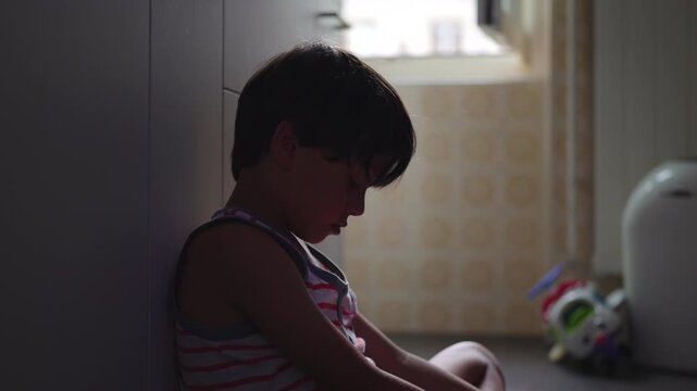 Young boy sitting against kitchen cabinet looking down with heavy expression after crying, quiet childhood moment expressing sadness reflection and emotional vulnerability