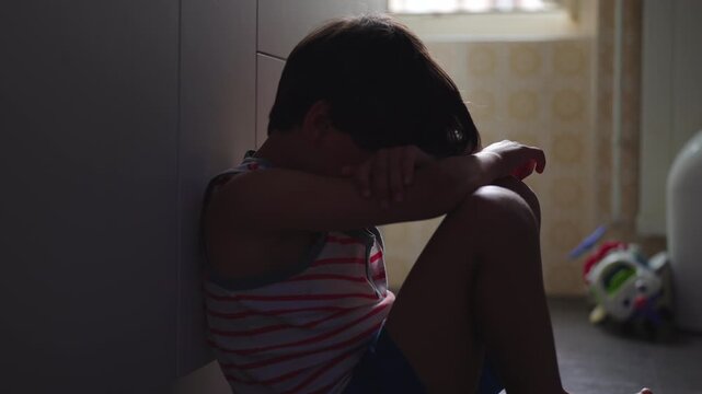 Young boy sitting on kitchen floor with arms covering face while crying quietly, emotional childhood moment expressing sadness vulnerability and distress at home