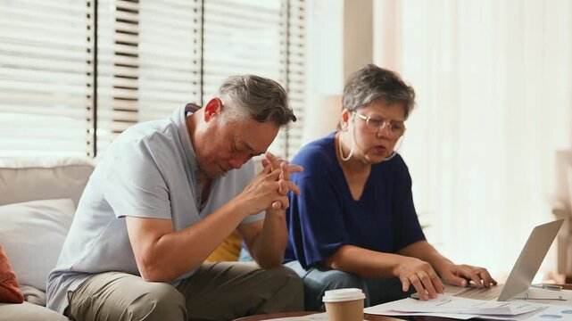 Unhappy Asian senior couple sitting on sofa at home facing financial problem, stress from debt and bills, worried about insurance and money, retirement struggle, emotional tension and family hardship.