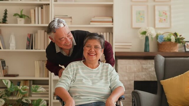 Portrait of elderly Asian husband supporting wife in wheelchair, both looking at camera with happiness, showing love, bonding, caregiving, strong relationship, family connection and warm retirement li