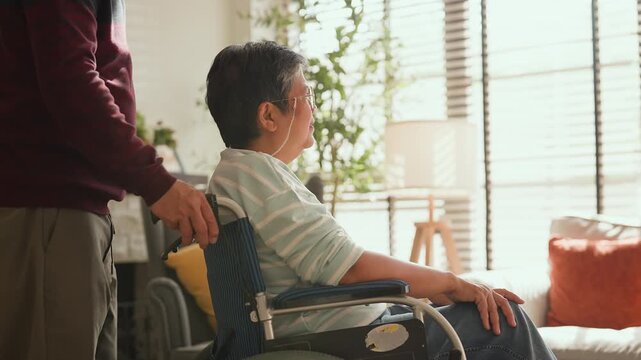 Asian senior couple at home, retired man in wheelchair embraced by loving woman, caregiving support, bonding and true love, warm living room lifestyle, family connection, compassion and lifelong partn