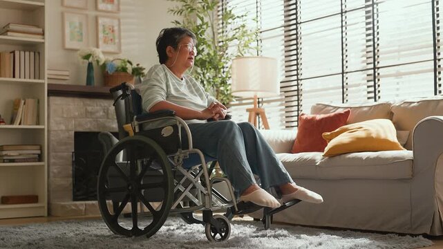 Elderly Asian woman in wheelchair looking out in living room, feeling lonely and missing freedom, retirement lifestyle change, emotional moment needing support, care, encouragement and family connecti