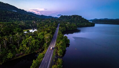 Obraz premium Aerial view of a car driving along a scenic road through lush tropical jungle landscape, calm lake reflection, twilight hour travel atmosphere