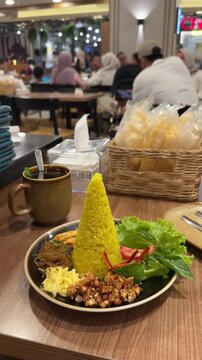 a plate of nasi tumpeng mini with a tall cone of yellow rice, surrounded by noodles, peanuts, egg, shredded chicken, and fresh greens. served with a cup of teh pandan jawa or javanese pandan tea