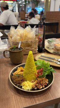 a plate of nasi tumpeng mini with a tall cone of yellow rice, surrounded by noodles, peanuts, egg, shredded chicken, and fresh greens. served with a cup of teh pandan jawa or javanese pandan tea
