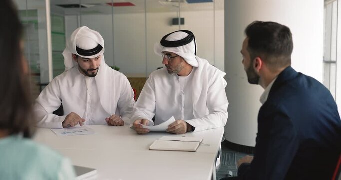 Senior and young Arabic businessmen analyzing paper marketing reports on meeting with European business partners, discussing partnership terms, getting agreement, shaking hands