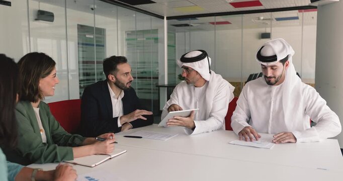Middle Eastern and Caucasian business partners discussing investment for online product, watching Internet content on tablet, signing contract, shaking hands at meeting negotiation table