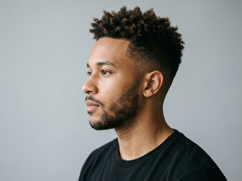 Young man with stylish afro fade haircut and beard, looking thoughtfully to the side against a soft gray background