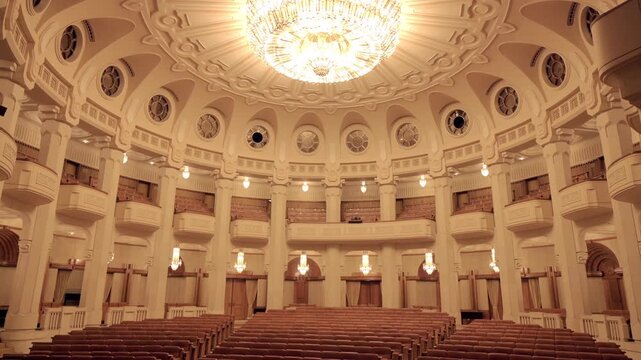 Elegant, empty hall with ornate ceiling, shimmering chandelier, and rows of chairs for grand events.