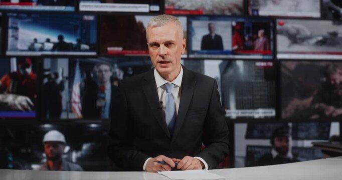 Focused Mature Man Reporter Sitting at White Desk, Talking to Camera. Concept of Massive Information Flow and Reporting on Casualties, Military Operations, and Diplomatic Summits in TV Studio.