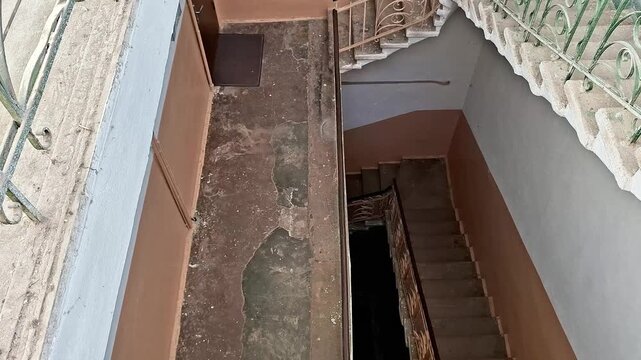 View from above of stairwell with green railings, beige walls, worn stone floor, and door on landing, highlighting aged textures, patterns, and quiet atmosphere of the space