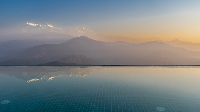 Dawn over the mountains. Beautiful view from the roof of the hotel. Snow-capped peaks against a blue sky illuminated by gold. Mirror image on the infinity pool water. Reflection. Nepal. Sarangkot.  

