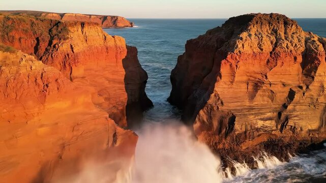Golden hour drone view of rust red coastal cliffs split by a narrow ocean channel, waves surging at the base of sculpted headlands in a dramatic remote shoreline landscape of raw geology