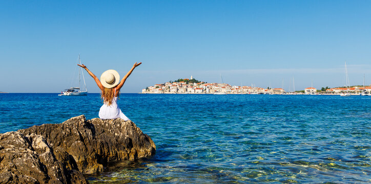 Happy Young Woman in White Dress and Sun Hat Sitting on Rocks Overlooking Primo&scaron;ten Old Town, Croatia