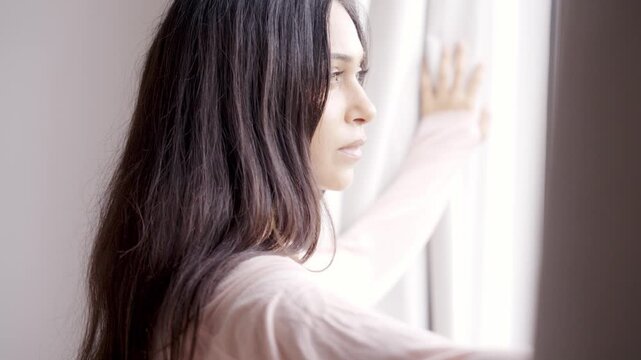 Attractive Young Woman Opening Window Curtains to Let Bright Morning Sunlight Enter Room. Beautiful Girl Looking Outside With Hope. One Adult Female Starting Her Day in Cozy Modern Residence 4k