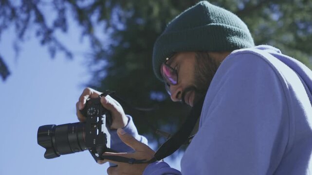 A young man with glasses standing in a park reviewing photos he took with a film camera. He is wearing a blue winter cloth in India. 