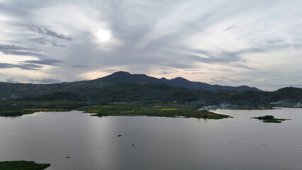 Fototapeta premium Tranquil Mountain Lake Landscape with Rural Village and Lush Green Hills Under a Bright, Cloudy Sky at Dawn