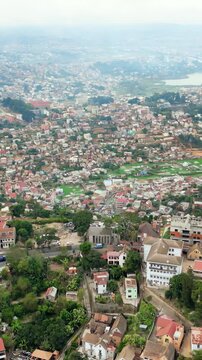 Aerial drone view above Antananarivo with the city skyline spread across surrounding hills, with dense urban neighborhoods and large historic palace complex overlooking Madagascar's capital. Vertical
