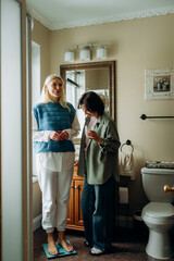 Fototapeta premium Two women stand together in a bright bathroom. One stands on a scale, while the other watches, holding a cup. Their expressions show support and encouragement as they share this health-focused moment.