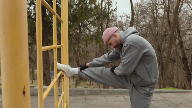 Bearded man stretching his leg on yellow metal ladder at outdoor fitness area. Male athlete in pink beanie warming up before street workout session in park