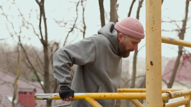 Close up of male athlete performing intense dips on parallel bars. Focused man in grey hoodie training on outdoor sports ground during autumn day