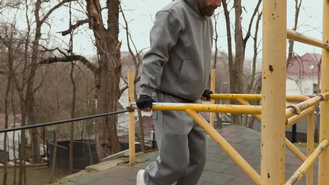 Side view of man performing dips on parallel bars at outdoor sports ground. Bearded athlete training triceps and chest during morning street workout