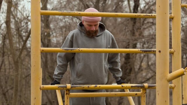 Front view of bearded man in pink beanie doing push ups on parallel bars. Sporty male in grey hoodie performing strength exercises on outdoor fitness ground