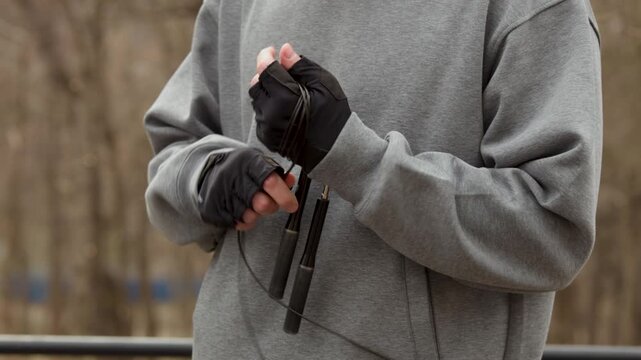 Close up of hands in fingerless gloves winding a jump rope after a workout. Athlete's hands packing sports equipment on an outdoor fitness ground