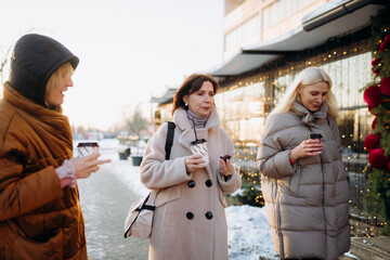 Obraz premium Three friends stroll down a snowy street, each holding a warm beverage. They share smiles and laughter, enjoying the winter sunlight and festive decorations around them.