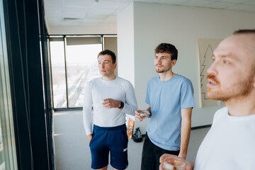 Fototapeta premium Three friends stand together inside a bright office, gazing out the window. They hold drinks in their hands, sharing a calm moment. Sunlight fills the space as they connect and reflect.
