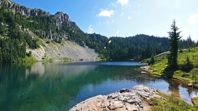 high-altitude view captures the brilliant turquoise waters of Lake Valhalla nestled in a rugged glacial cirque, surrounded by dense evergreen forests and steep mountain slopes under a bright blue sky 