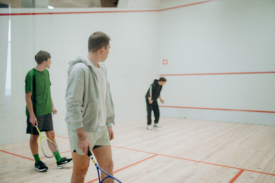 Three friends gather in a well-lit squash court, each holding a racket. They prepare for an exciting match filled with skill and camaraderie. Laughter fills the air as they play.