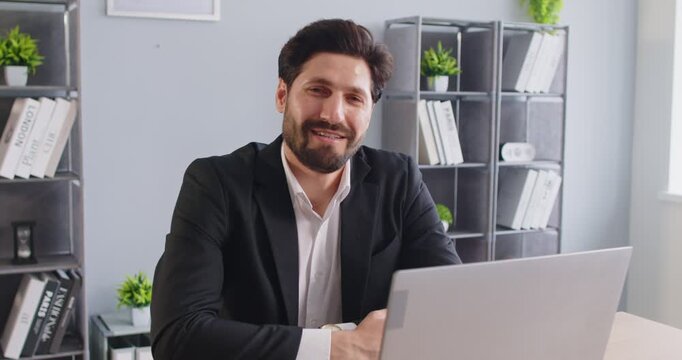 Busy confident businessman working on laptop at office desk. Young man with beard and suit looking at camera during break from work and smiling, happy professional entrepreneur sitting at workplace