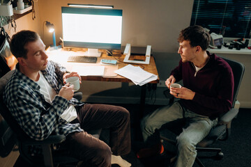 Two friends enjoy warm cups of coffee while engaged in a deep conversation. The dimly lit room features a computer, scattered papers, and a relaxed atmosphere, perfect for sharing ideas.