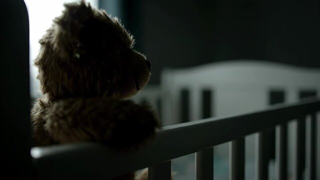 Teddy bear in white crib. Childhood toy in dark room. Loneliness concept.