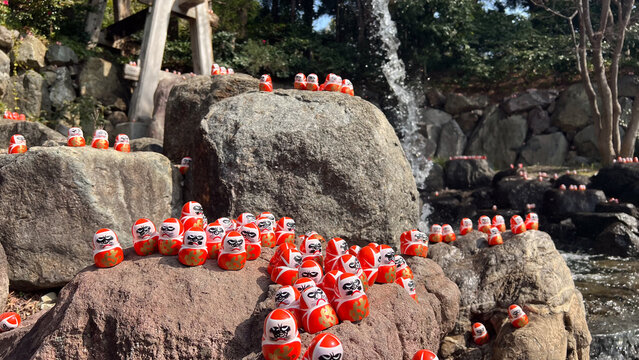 大阪箕面市の勝尾寺にある庭園の岩場に並んだ小さな勝ち運だるまたち、清らかな噴水の水しぶきと自然の岩が織りなす境内の風景、日本の伝統的な縁起物と自然が調和した癒やしの観光風景、高品質な旅行広告用風景写真。