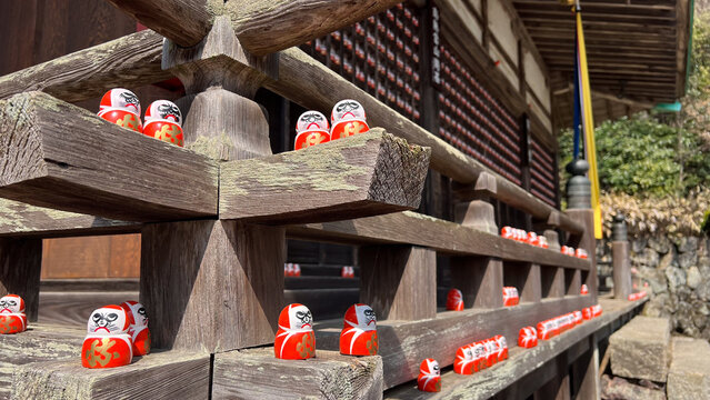 大阪箕面市の勝尾寺にある古い木造建築の欄干に置かれた小さな勝ち運だるまたち、歴史を感じさせる境内の静寂と伝統的な日本文化の風景、おみくじの後のダルマを奉納する習慣を捉えた高品質な文化観光写真。