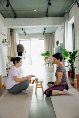 Fototapeta premium Two women sit cross-legged on soft mats, focusing on a small table with calming items. Natural light fills the peaceful studio, creating a relaxing atmosphere for their meditation.
